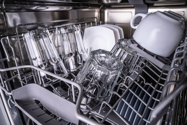 Egy tiszta tál mosogatógépben. Ragyogó átlátszó szemüveg és fehér csésze mosogatógépben történő mosás után. - Fotó, kép