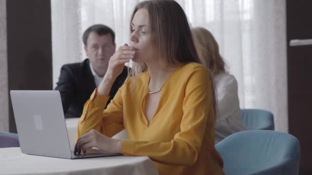 Confident young Caucasian woman yawning, closing laptop and moving device aside. Portrait of exhausted charming businesswoman overworking. Colleagues talking at the background in office. - Felvétel, videó