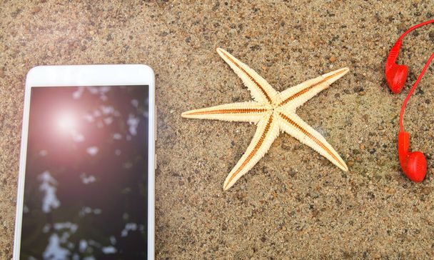 Strand nyaralás és utazás koncepció, okostelefon, kamera, fejhallgató és tengeri csillag a homokban a strandon, napfény tükröződik az okostelefon képernyőjén. - Fotó, kép