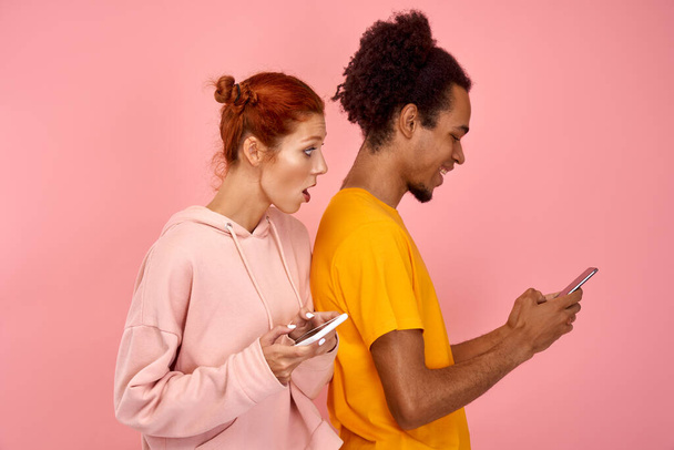 Suspicious jealous puzzled wife wants to spy sms message on husbands device, suspects of betrayal, looks over back, has embarrassed gaze. Young couple use modern gadgets for online communication - Photo, Image
