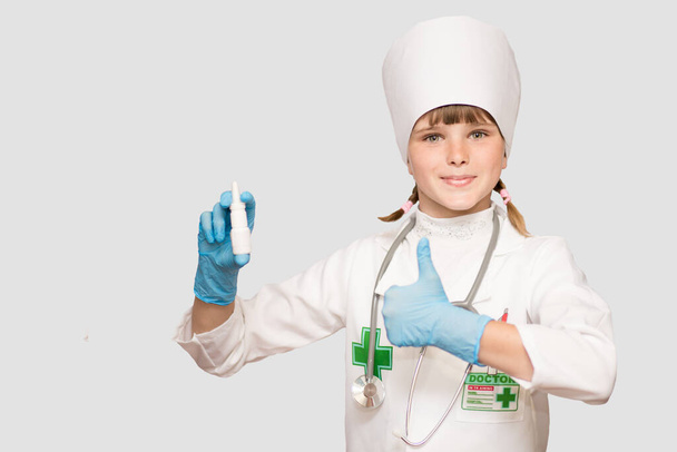 Mosolygó kislány orvosi egyenruhában, orrspray-vel vagy cseppekkel az egészségéért fehér üvegben. Orvos lány javasolja gyógyszer orrspray - Fotó, kép