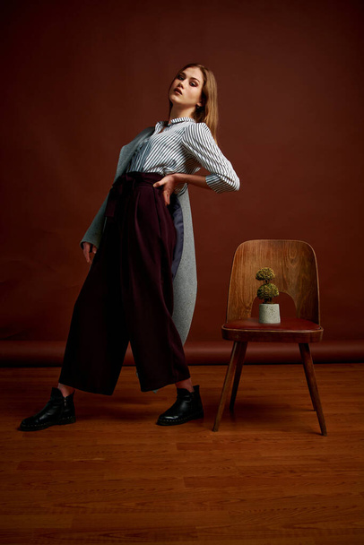 Mininalismi tyyli klassinen studio valokuva joukko nuori itsenäinen nainen. Upea nuori nainen klassisessa puvussa, mustat kengät ja harmaa takki. Vintage-tuoli. Naisen vaatteet. Muodikas vaatteita
. - Valokuva, kuva