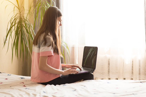 mladá krásná žena na posteli v blízkosti okna pomocí notebooku doma. Práce na dálku. Úkol. Zůstaň doma - Fotografie, Obrázek