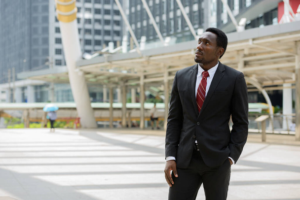 Портрет молодого привлекательного африканского бизнесмена в костюме против вида современного здания в городе на открытом воздухе - Фото, изображение