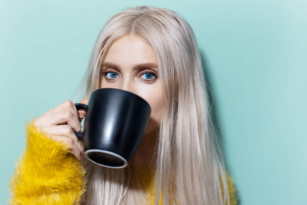 Studio portrait of beautiful blonde girl with blue eyes drinking tea from black cup, wearing yellow sweater on cyan, aqua menthe color. - Foto, imagen