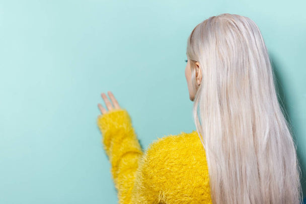 Back view of blonde girl wearing yellow sweater, touching studio background of cyan, aqua menthe color with copy space. - Foto, Imagen