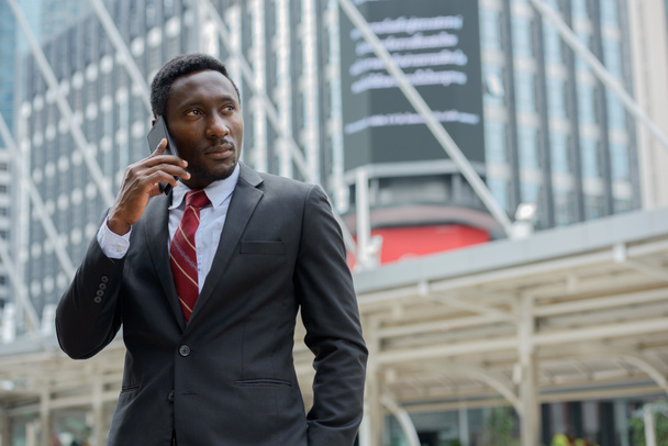 Portrait of young handsome African businessman in suit against view of modern building in the city outdoors - 写真・画像
