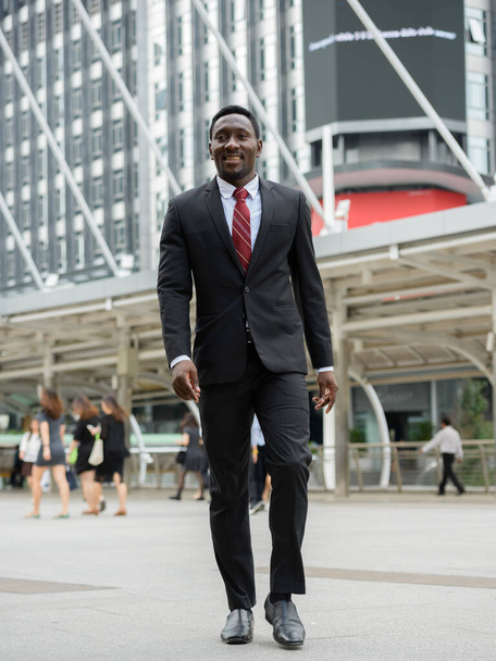 Portrait of young handsome African businessman in suit against view of modern building in the city outdoors - Fotó, kép