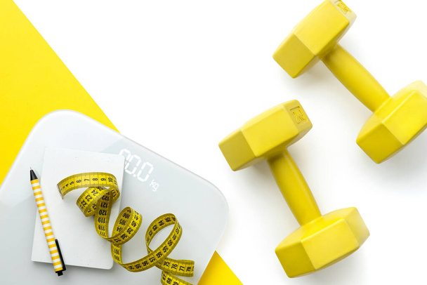 Flat lay top view scales, dumbbell and tape measure on white yellow double background with copy space. Personal fitness program workout concept - Photo, Image