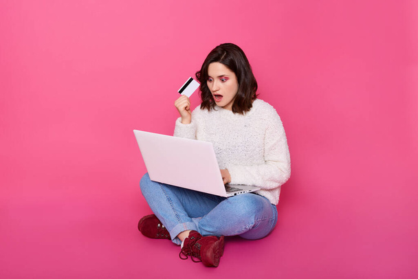 Indoor shot of woman sitting on floor, wearing casual attire, having astonished facial expression, keeps mouth opened, holding credit card in hand, doing online shopping, being shocked from prices. - Foto, Imagem