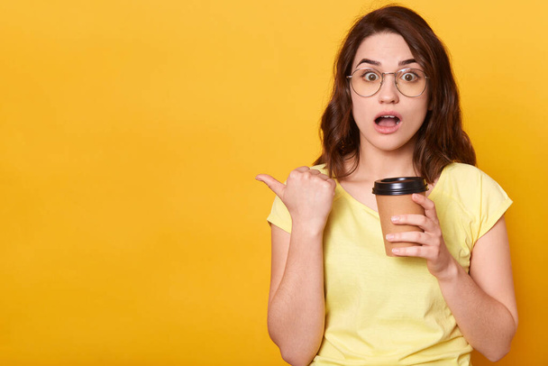 Closeup portrait of astonished woman posing against yellow studio background, looks surprised, being shocked, holding takeaway coffee, pointing aside, keeps mouth opened. Copy space for advertisement. - Foto, Imagen