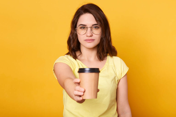 Closeup portrait of brunette girl offers takeaway coffee to somebody, lady wearing casual t shirt and spectacles, posing isolated on yellow studio background, attractive female looks calm and serious. - Fotografie, Obrázek