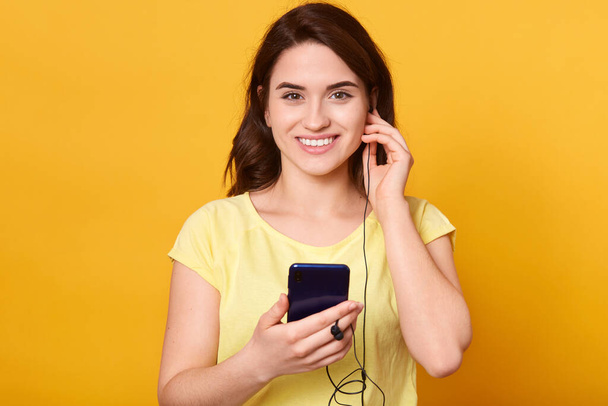 Studio shot of smiling girl with dark hair and pleasant appearance, looking directly at camera, wearing casual t shirt, listening to music indoors, spending her spare time. People hobby concept. - Photo, image