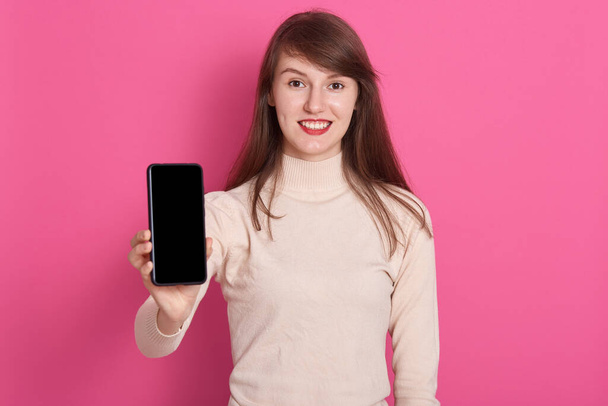 Horizontal shot of happy smiling female wearing casual shirt, holding modern smart phone with blank screen, showing cellphone with copy space for advertisement, stands isolated over pink background. - Fotoğraf, Görsel