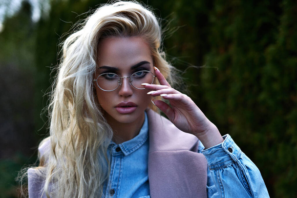 Young pretty blonde girl walking outdoor in winter cloth using thuja like background and posing for camera with sunglasses with full makeup and eyes like her blue jeans jacket - Fotoğraf, Görsel