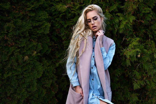 Young pretty blonde girl walking outdoor in winter cloth using thuja like background and posing for camera with sunglasses with full makeup and eyes like her blue jeans jacket - Foto, afbeelding