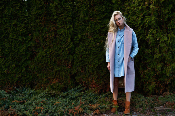 Young pretty blonde girl walking outdoor in winter cloth using thuja like background and posing for camera with sunglasses with full makeup and eyes like her blue jeans jacket - Foto, immagini