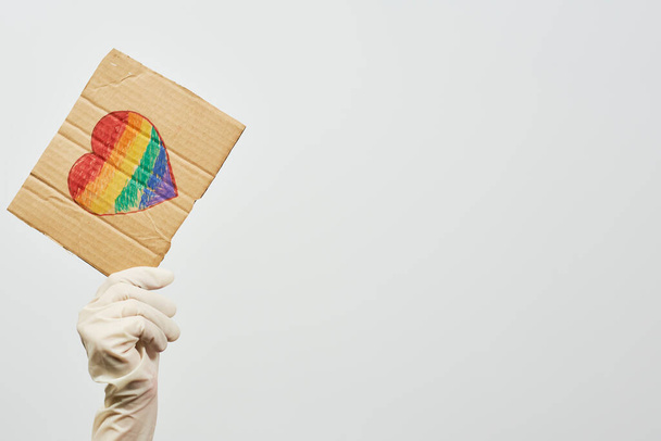 LGBTQ-freundliche Mitarbeiterin des Gesundheitssystems mit einem handgefertigten Plakat mit herzförmiger Regenbogenfahne. LGBTQ-Arzt kämpft an vorderster Front gegen Coronavirus - Foto, Bild
