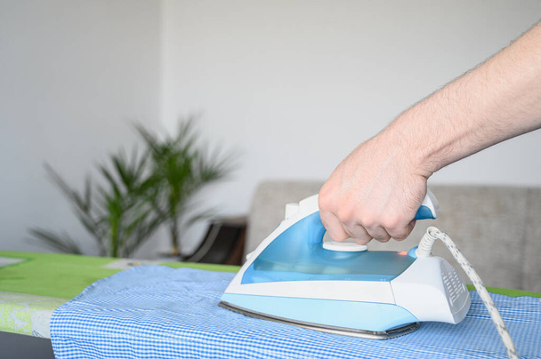 A férfi kéz vasat tart. Rutin házi feladat. Egy kék kockás inget lóbálni egy vasalódeszkán egy fényes szobában.. - Fotó, kép