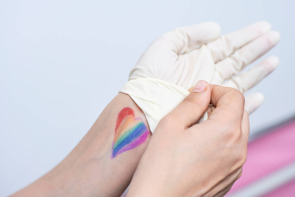 Une femme médecin LGBTQ se bat avec le coronavirus en première ligne. Travailleur du système de santé LGBTQ amical avec drapeau arc-en-ciel au poignet. LGBTQ médecin amical ou fournisseur de soins de santé enlève des gants chirurgicaux
 - Photo, image