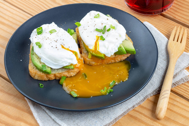 Toasts with poached eggs and slices of avocado on a black plate (on napkin on wooden background). Tasty, quick and healthy breakfast. - Foto, immagini