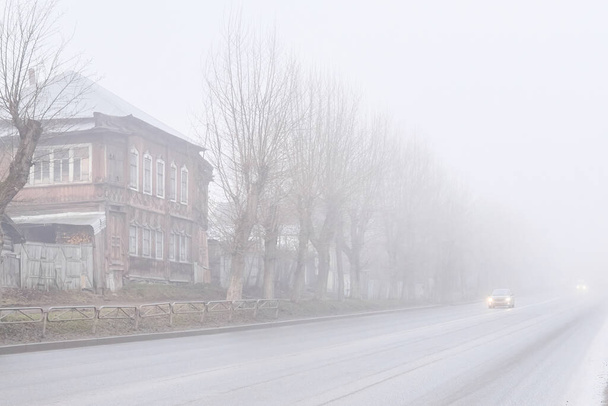 сельская асфальтовая дорога с деревянными домами и придорожными ограждениями, скрывающимися в мутном утреннем тумане, видна движущаяся к ней машина
 - Фото, изображение