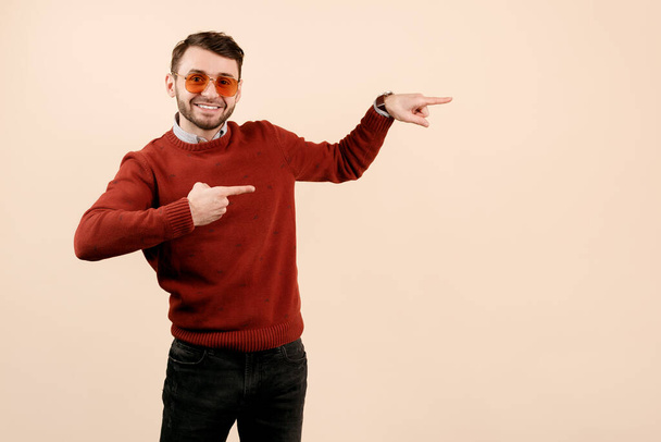Cheerful bearded man in casual outfit wearing sunglasses pointing fingers away on beige background. - Foto, Imagem