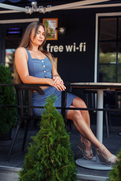 Függőleges fotó egy lányról egy kávézóban. Egy kék felsőben ülő lány oldalra ül. Wi-Fi zóna. Zöld thuja a kávézóban. - Fotó, kép