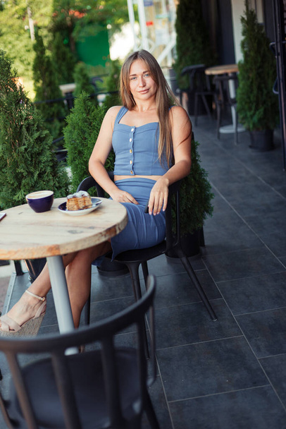 Vertical photo of a girl. Attractive girl in a blue top sits in a European cafe. Green thujas. - Φωτογραφία, εικόνα