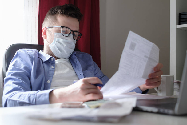 Young caucasian man in medical mask working with laptop at home browsing bills and documents. Businessman going through paperwork at home office. Stay home and self isolation quarantine. - Photo, Image