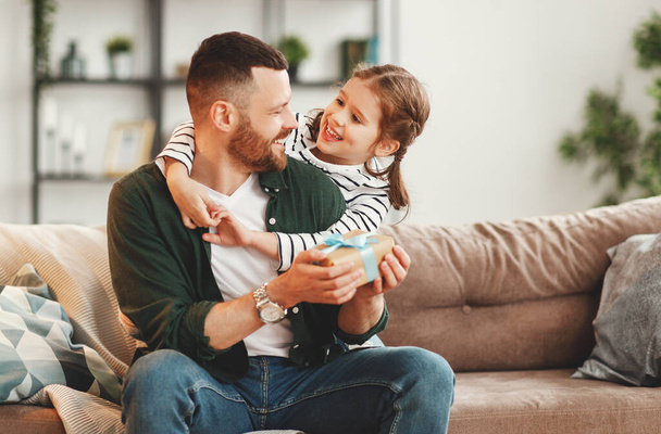 Happy young man with gift in hand and adorable little daughter embracing and   looking at each other while sitting together on sofa in cozy living room on fathers da - Photo, Image