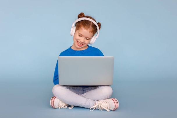 Pełne ciało pozytywna dziewczynka w słuchawkach uśmiecha się i ogląda kreskówkę na nowoczesnym laptopie siedząc skrzyżowane nogi na niebieskim tle - Zdjęcie, obraz