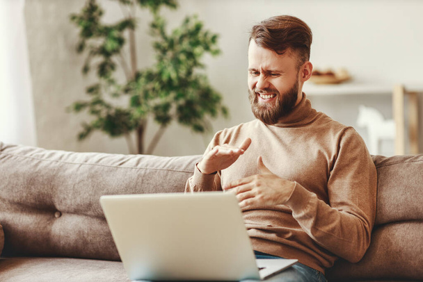 Позитивний молодий чоловік в повсякденному одязі сидить на дивані і спілкується з другом під час відеодзвінків на ноутбук, проводячи час вдома
 - Фото, зображення