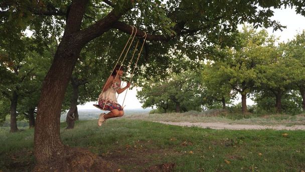 オークの木の枝で揺れるロープの上で揺れる自由な少女。太陽の下で公園でスイングする健康な子供。女の子は森の中でスイングに飛ぶのが大好きです。幸せな家族や子供時代の概念は. - 写真・画像