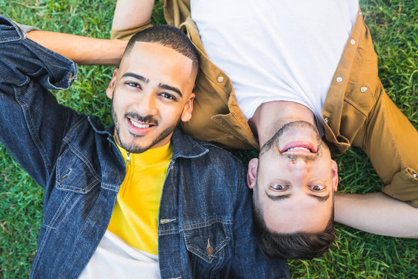 Portrait of young gay couple spending lovely time and laying down on the grass at the park. Lgbt and love concept. - Фото, изображение