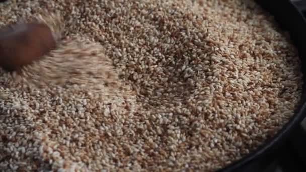 Zbliżenie nasion sezamu smażonych na patelni, dłoń samicy miesza nasiona sezamu z drewnianą łyżką, selektywne skupienie - Materiał filmowy, wideo