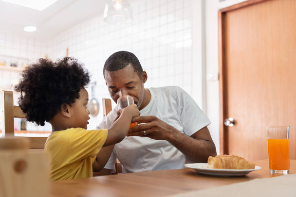 Happy African American family enjoying with drinking orange juice in dining room. Joyful Black Man in white cloth and little kid boy in yellow shirt with breakfast or brunch. Lifestyle. - Foto, Imagem