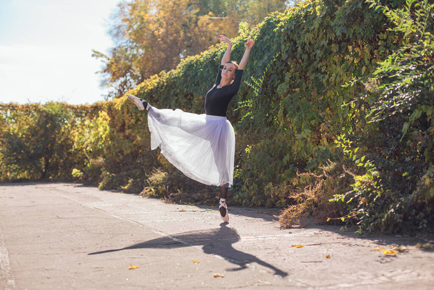 Woman ballerina in a white ballet skirt dancing in pointe shoes in a golden autumn park. Ballerina standing in beautiful ballet pose - Фото, изображение