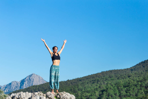 Meisje beoefent yoga op de top van de berg, vrouw voert asanas staande op een grote steen tegen een achtergrond van bergen en een hemel, Meditatie in de natuur, Oefening voor de ontwikkeling van evenwicht. - Foto, afbeelding