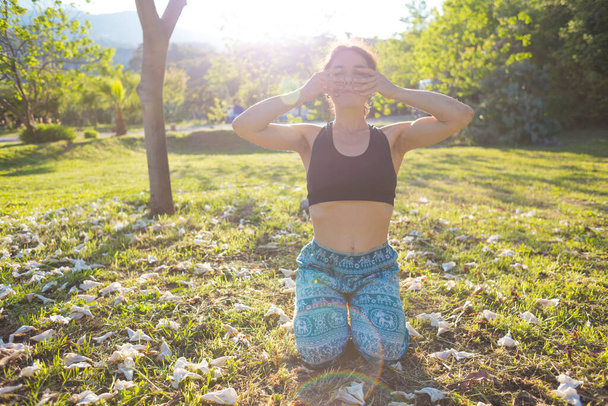 Женщина делает самомассаж лица, девушка практикует йогу на зеленой траве на фоне зеленых деревьев, женщина выполняет асаны на закате, медитация на природе, женские пальцы на лице. - Фото, изображение