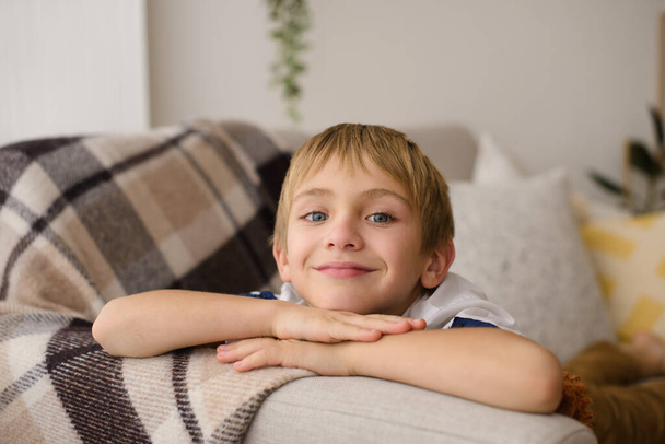 πορτρέτο του όμορφου μικρού αγοριού αναπαύεται στο στομάχι του στον καναπέ, με καρό καρό καρό, στο δωμάτιο στο σπίτι - Φωτογραφία, εικόνα
