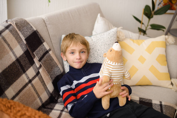 χαμογελαστό αγόρι ξαπλώνει σε γκρι καναπέ, κρατά στα χέρια του παιχνίδι αρκουδάκι, το πρωί στο σπίτι στο δωμάτιο - Φωτογραφία, εικόνα