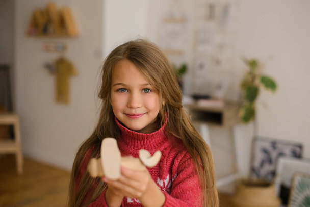 girl with long hair in pink sweater in white room at home, plays wooden toy airplane - Zdjęcie, obraz