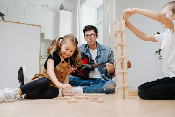 Enthusiastic dad playing guitar on the kitchen floor, watching his little son and daughter playing with wooden blocks. Building tower. Spending time with kids on isolation. - Foto, imagen