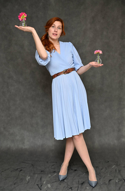The model demonstrates standing in full growth different poses with a flower in her hands. Vertical portrait of a young pretty red-haired woman in a blue dress on a gray background. - Foto, imagen