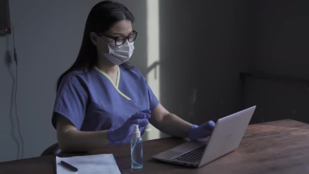 Νοσοκόμα με ιατρική στολή και γάντια μιας χρήσης σκουπίζει το laptop της πριν το χρησιμοποιήσει κατά τη διάρκεια της περιόδου πανδημίας καραντίνας. Πανδημία του Coronavirus. Πρότυπα 422 - Πλάνα, βίντεο