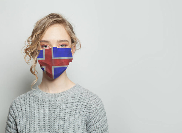 İzlanda bayrağı taşıyan yüz maskesi takan genç bir kadın. Grip salgını ve virüs koruma kavramı - Fotoğraf, Görsel