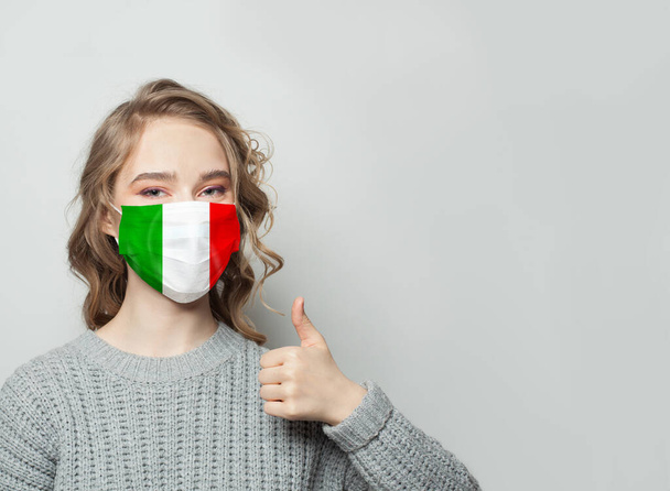 Mujer feliz con máscara facial sosteniendo el pulgar hacia arriba con fondo de bandera nacional italiana. Epidemia de gripe y concepto de protección del virus
 - Foto, Imagen