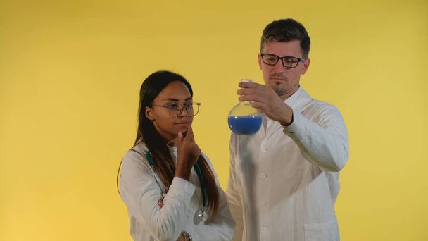 Laboratuvar önlüklü kadın ve erkek, sarı arka planda deneysel sıvı bulunan mataraya bakıyor.. - Fotoğraf, Görsel