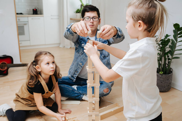 Papa speelt met kinderen op de keukenvloer, helpt zijn zoontje en dochter met het bouwen van een toren van houten blokken. Tijd doorbrengen met kinderen op isolatie. - Foto, afbeelding